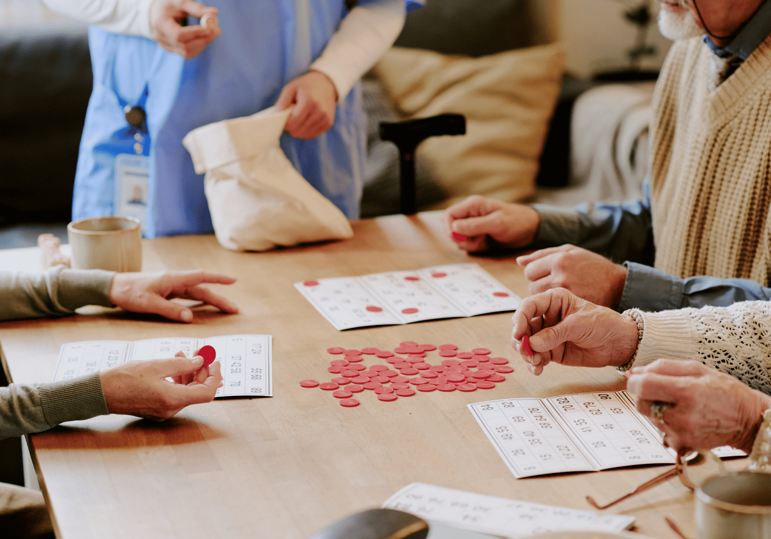 Group of seniors playing bingo at a table with red tokens and numbered cards; a caregiver stands in the background.