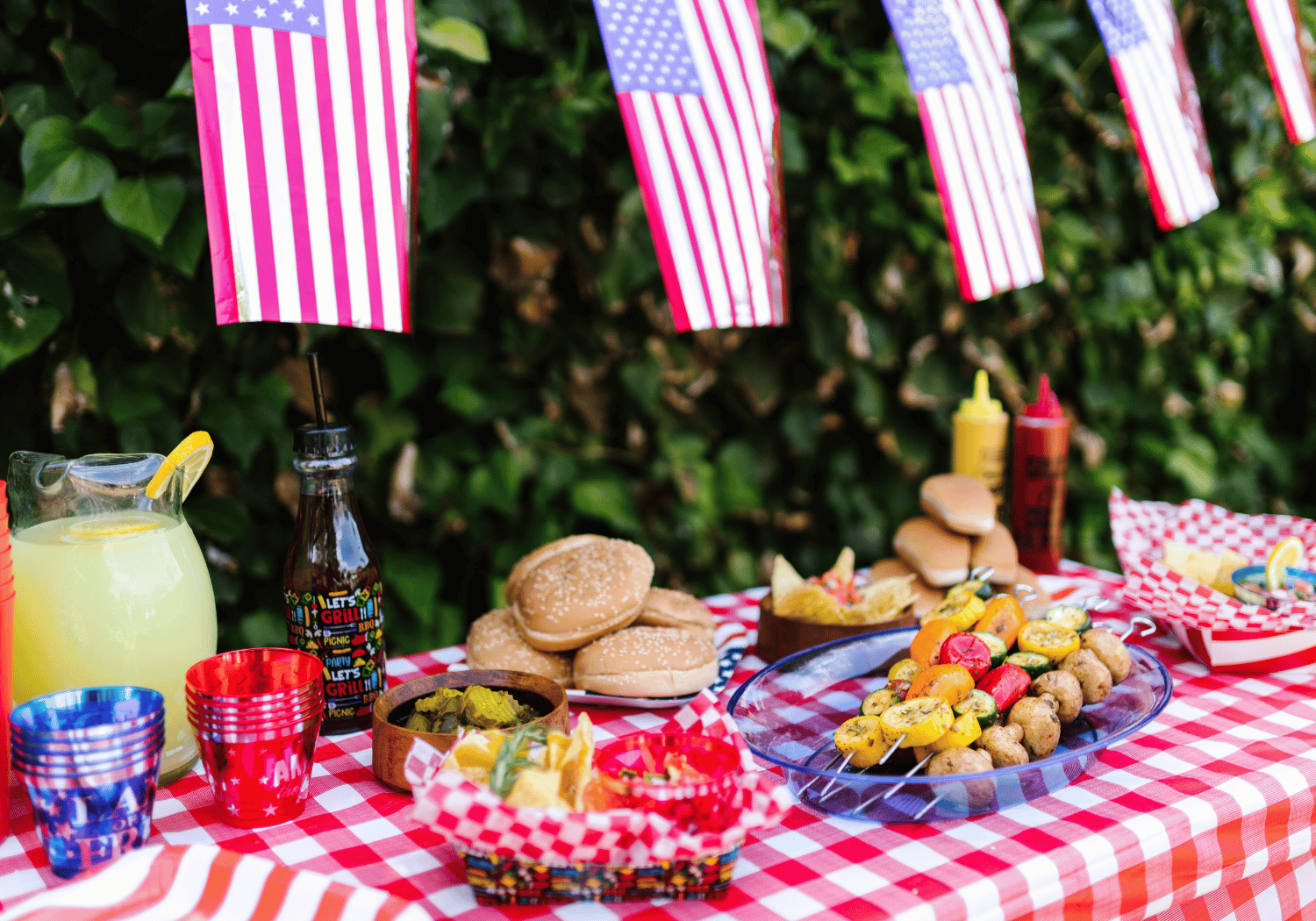 Patriotic picnic scene with American flags, burgers, veggie and potato skewers, chips, condiments, and lemonade on a red‑and‑white checkered tablecloth.