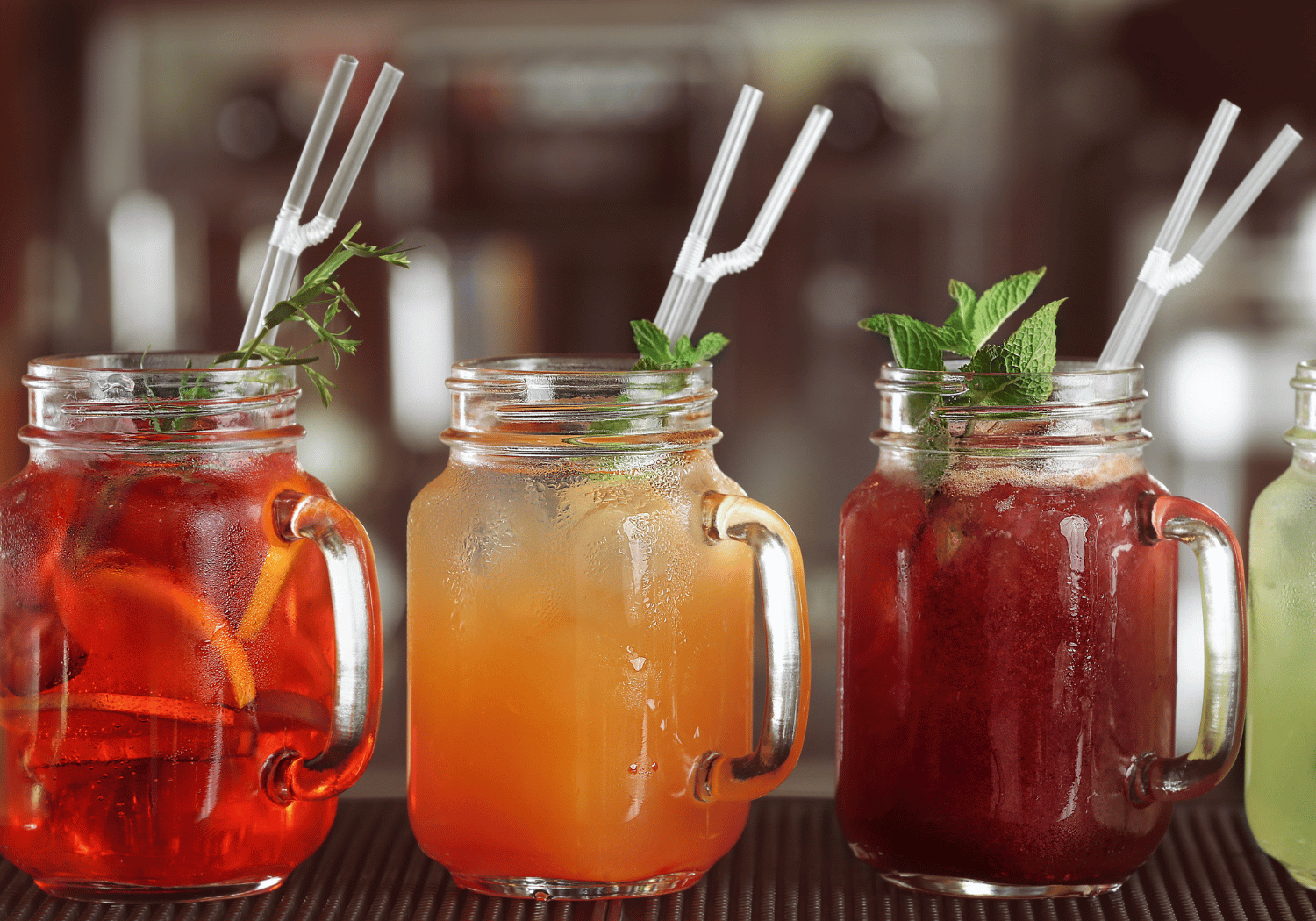 Three colorful iced drinks in mason jars with handles, each garnished with mint and a white striped straw on a bar counter.