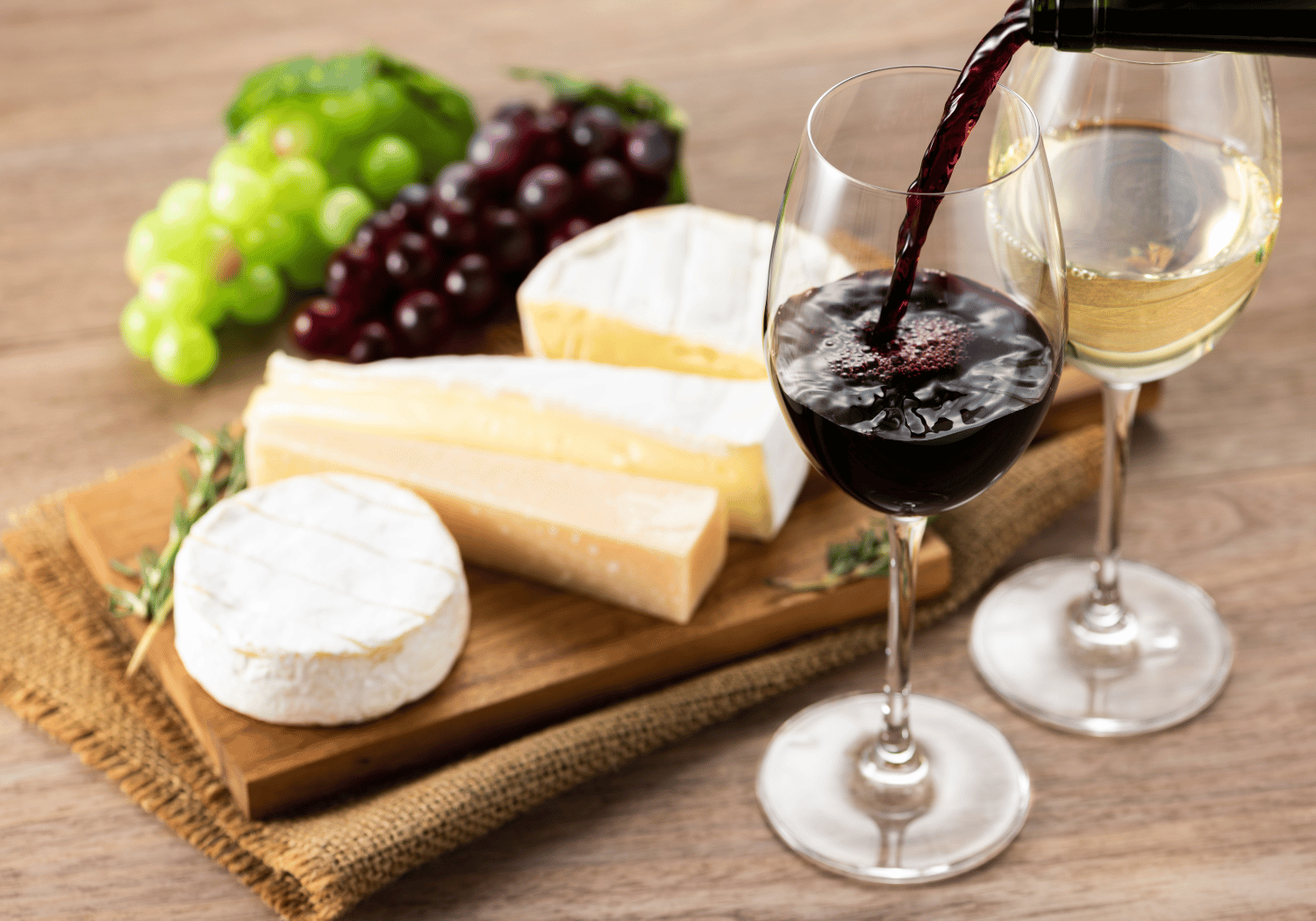 Wine is being poured into a glass beside a cheese board with Brie, other cheeses, and grapes on a wooden table.